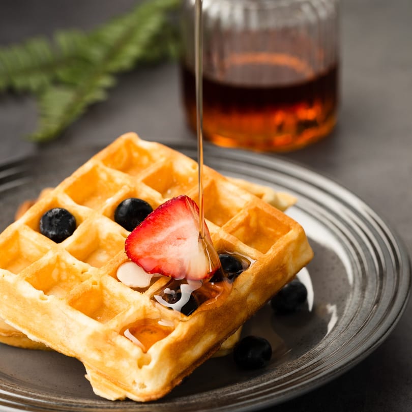 Golden waffles topped with strawberries, blueberries, and syrup pouring from above.
