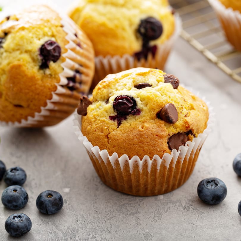 Three golden-brown blueberry and chocolate chip muffins in white paper liners, with loose blueberries scattered around.
